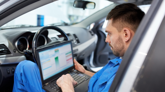 Male car mechanic apprentice sitting inside car with laptop