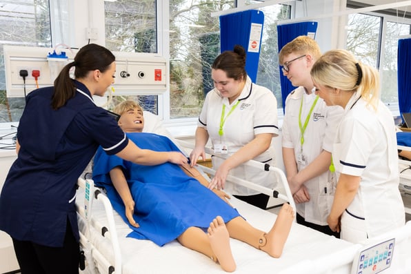 Healthcare students and teachers stand around a lifesize simulation doll in a hospital bed