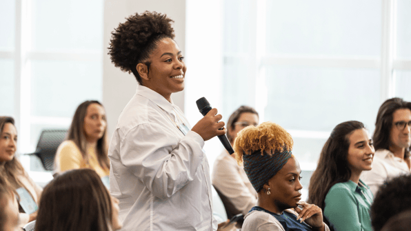 Young black female standing up from a seated crowd and speaking into a microphone