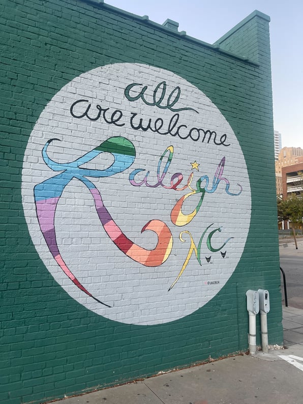 A mural on a green wall saying 'All are welcome Raleigh N.C'