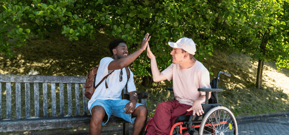 A young black man sitting on a park bench high-fiving a young white man in a wheel chair
