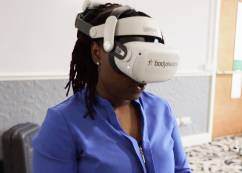 a female student with dark, medium length dreadlocks wearing a VR headset and a blue button down shirt  