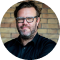 Round-cropped headshot Tom Wambek wearing glasses and a black shirt.