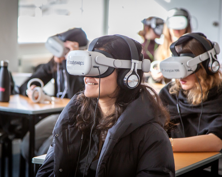 VR headsets are being used by a classroom of students in black hoodies sitting at desks.