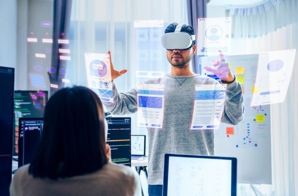 Asian software developer wearing a virtual reality headset works on a VR project, with a colleague focused on her computer